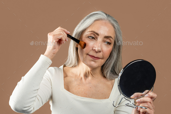 Smiling elderly caucasian woman with gray hair applying blush on cheeks, looks in mirror isolated on