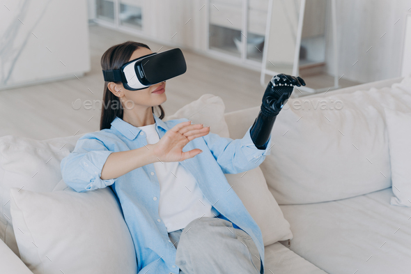Young disabled girl with bionic prosthesis arm, virtual reality glasses playing video game at home