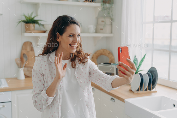 Young spanish woman has video call on phone at kitchen. Girl is waving hand and smiling. - Stock Photo - Images