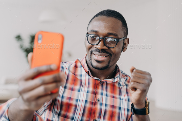 Happy african american man wearing glasses holding smartphone smiling reading good news message - Stock Photo - Images