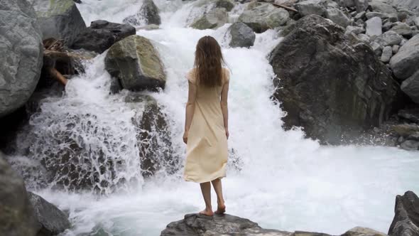 Young Happy Woman in Orange Dress Standing Under Spray of Water on Background of Big Waterfall