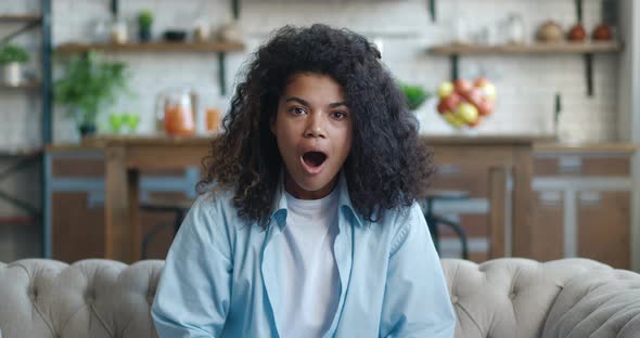 Close Up Portrait of Young African American Woman with Wow Expression While Sitting on Sofa at Home alt