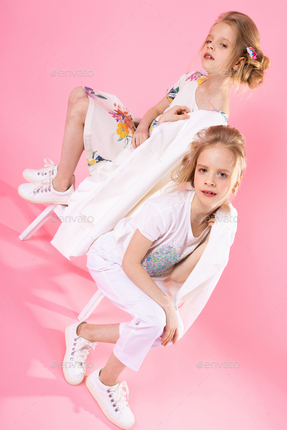 Twins girls in bright clothes posing near the stairs with two steps on a pink background.