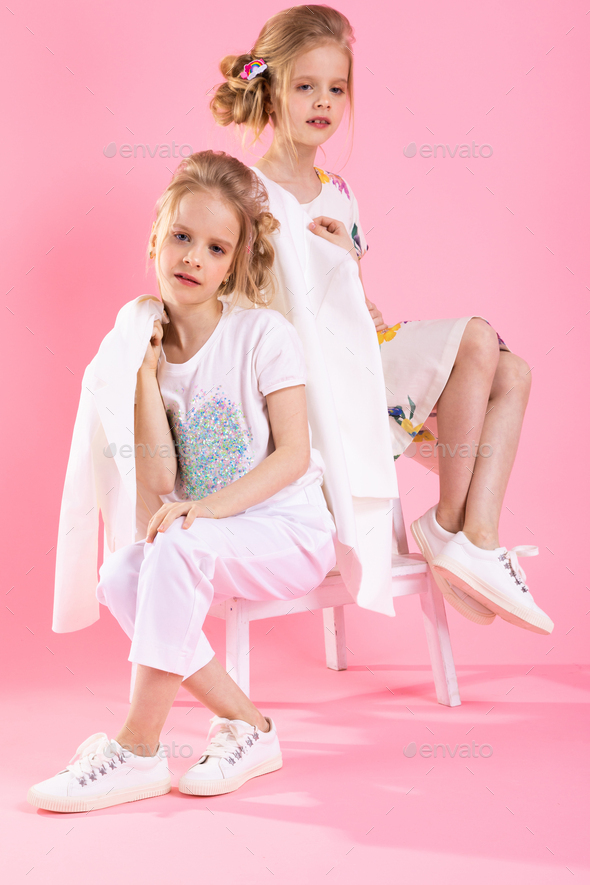 Twins girls in bright clothes posing near the stairs with two steps on a pink background.