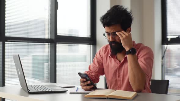 Cheerful Bearded Indian Businessman in Glasses Holding Mobile Phone Texting Online Message Sitting alt