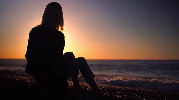 blonde in denim suit is sitting on seashore at sunset with smartphone in hands alt