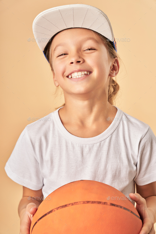 Cheerful female child in cap holding basketball ball - Stock Photo - Images