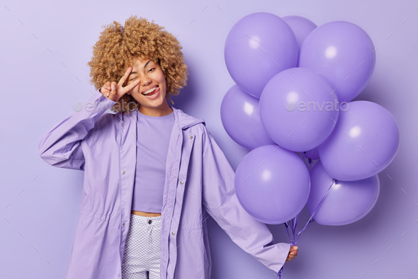 Horizontal shot of positive woman with curly hair makes peace sign holds inflated balloons gathered