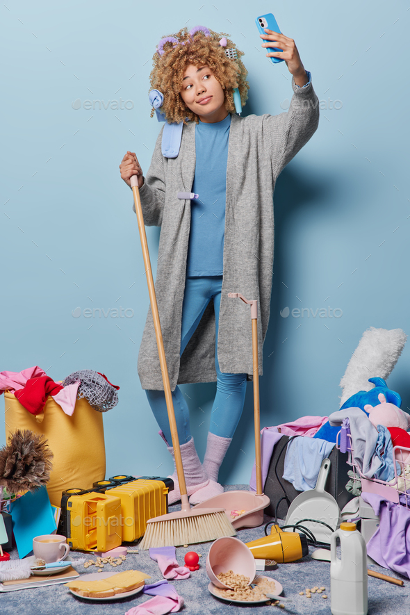 Vertical shot of curly haired housewife dressed in domestic clothes sweeps floor takes selfie via