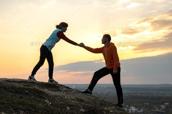 Man and woman hikers helping each other to climb stone at sunset in mountains.