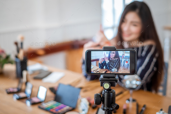 Young asian woman blogger with makeup cosmetics recording video clip online by smartphone at home.