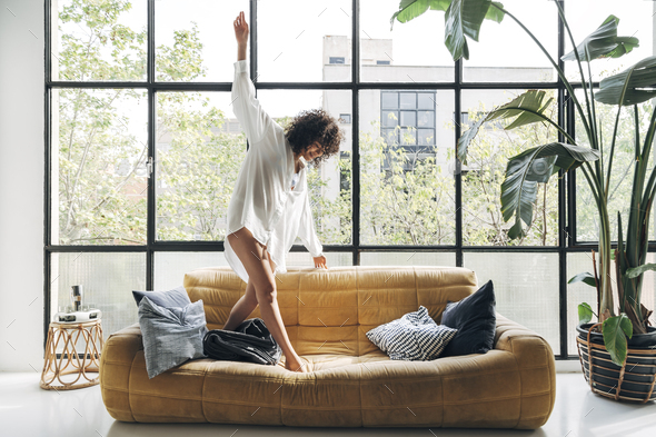 African american young woman dancing on couch, having fun, carefree in a bright loft apartment