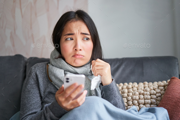 Asian girl with influenza, sits at home, cries and looks upset, holds smartphone, feels unwell and