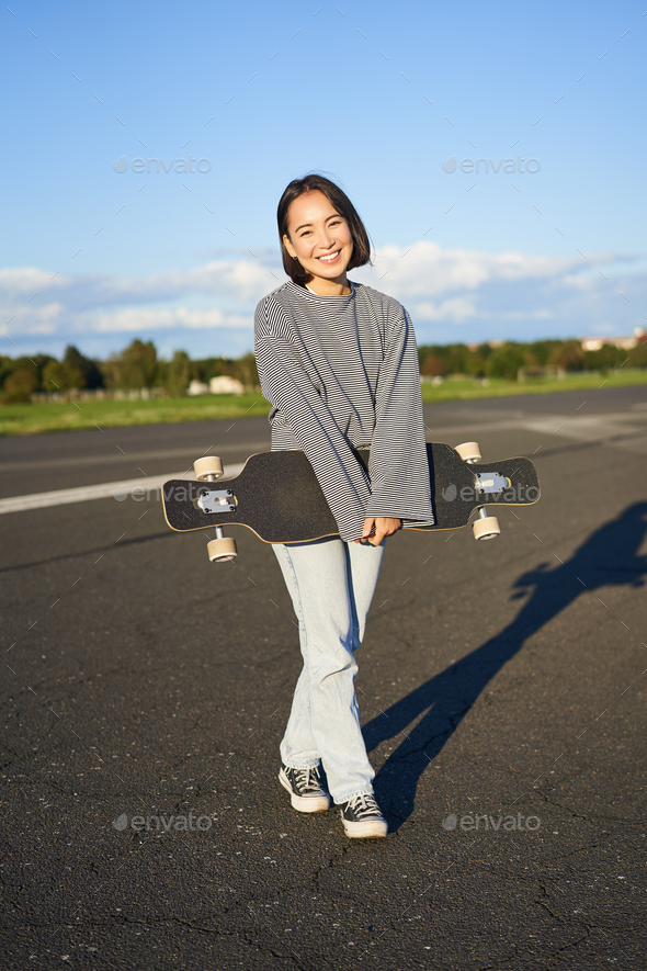 Vertical shot of skater girl posing with longboard, cruising on empty road in suburbs. Smiling