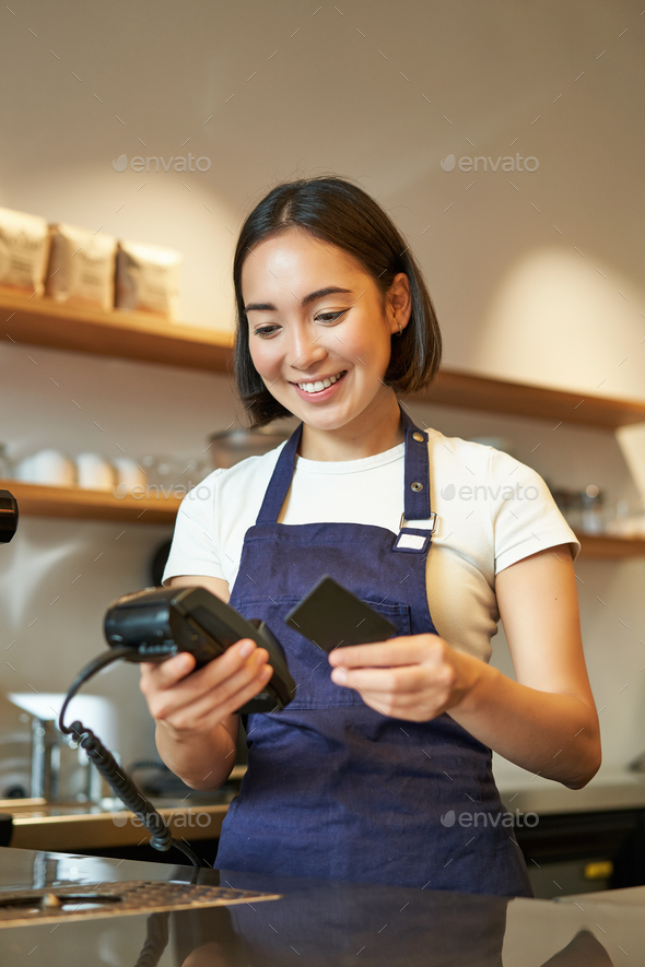Portrait of smiling asian barista, coffee shop employee using POS terminal and credit card, helps