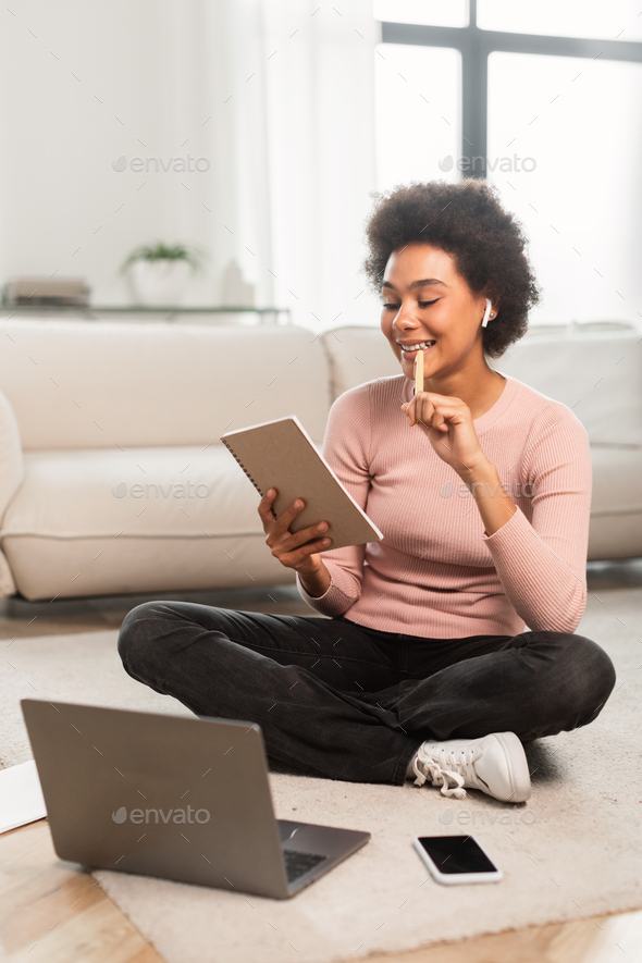 Thoughtful smiling young mixed race female with computer makes notes, plans work, sits on floor