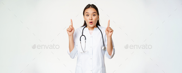 Portrait Of Surprised Female Asian Doctor Physician Pointing Fingers Up Showing Amazing News