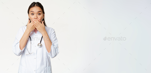 Image of shocked female asian doctor covers her lips with hands, close mouth and looking worried