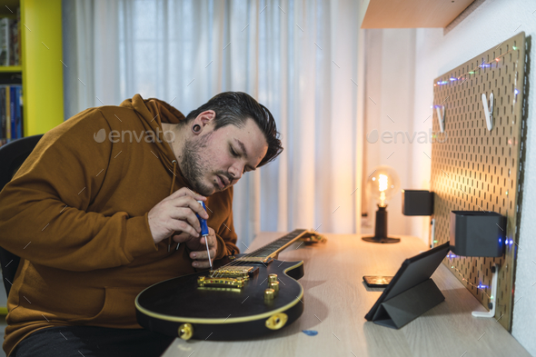 Selective focus shot of a guy with piercings wearing a brown hoodie repairing a guitar in a room
