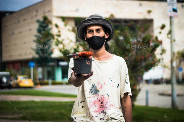 Handsome graffiti artist in a face mask and paint-stained clothes holding a paint can and brush