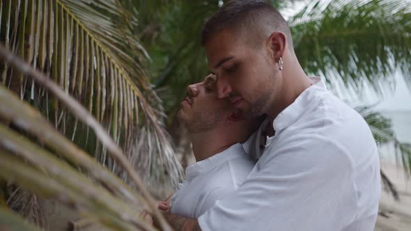 Gay Man Embracing His Partner From Behind Standing on a Tropical Beach By the Palm alt