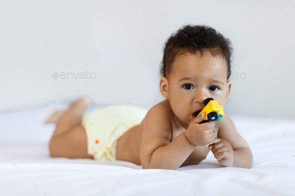 Cute African American Infant Baby Lying In Bed And Biting Toy - Stock Photo - Images