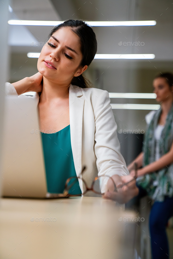 Young woman at office desk working on laptop with stress, neck pain and exhaustion.