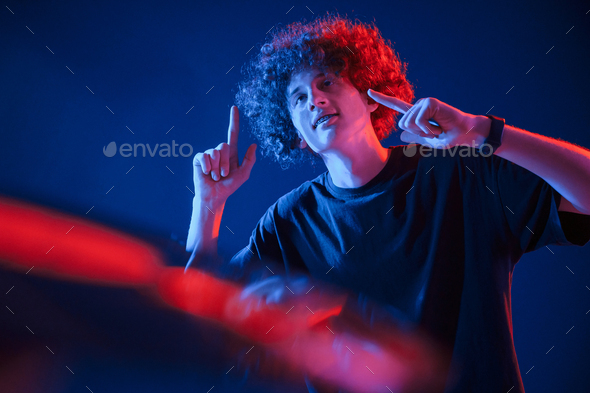 Dj is playing music from vinyl. Young man with curly hair is indoors illuminated by neon lighting