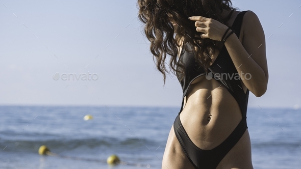 Curly female with belly button piercing in elegant black bikini posing at the beach