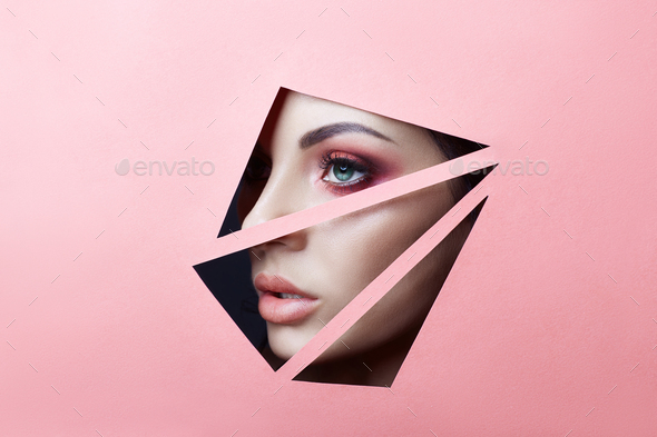 Beauty face red makeup eyes of a young girl in slit hole of pink paper. Woman with beautiful makeup