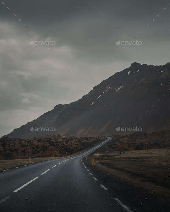 Empty curvy road next to a beautiful rocky mountain under a gray gloomy sky