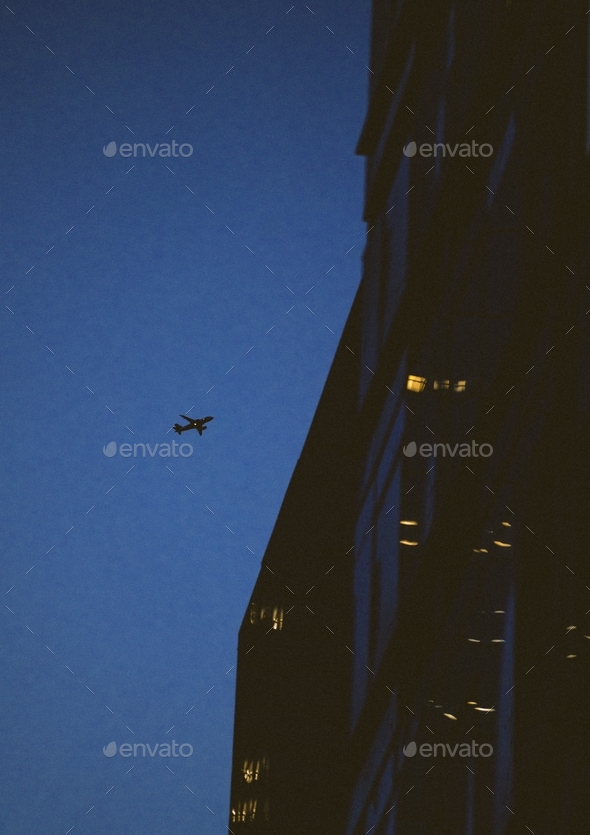 Silhouette of an airplane under the dark sky at night and apartment buildings with turned-on lights