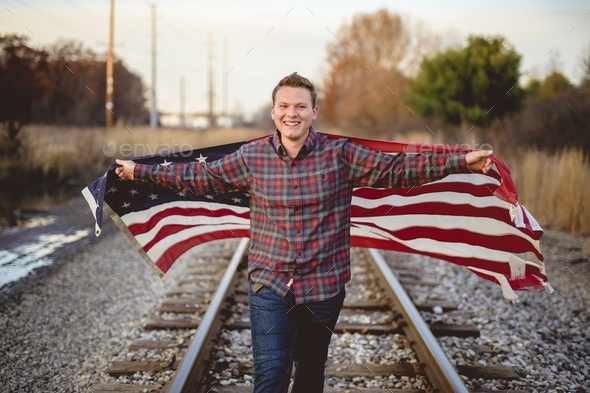 Smiling male holding the United States flag while walking on the train rails