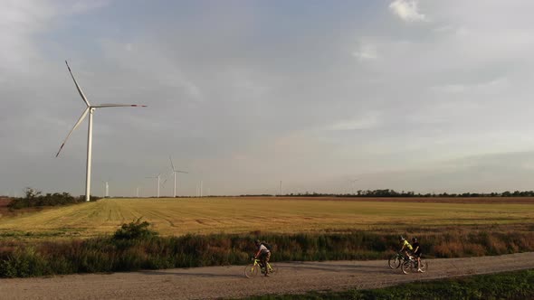 Bicyclists Ride Along Rural Road Past Field with Windmills