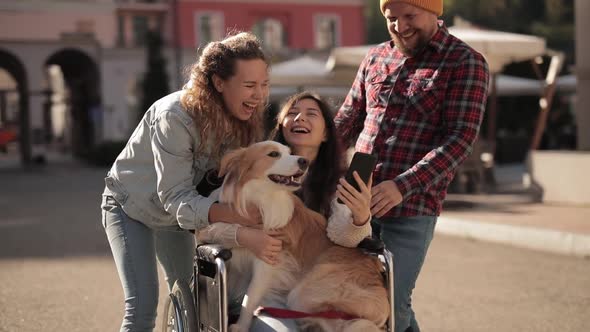 Disabled Invalid Girl Sitting on Wheelchair and Laughing with Friends Looking at Smartphone Outdoors alt