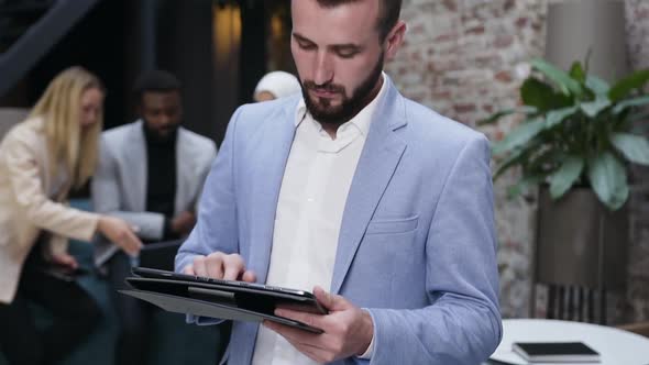 Businessman Making Notes in Tablet Computer App on the Background of His Co Workers alt