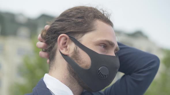 Close-up Side View of Young Caucasian Businessman in Covid-19 Face Mask Standing Outdoors alt