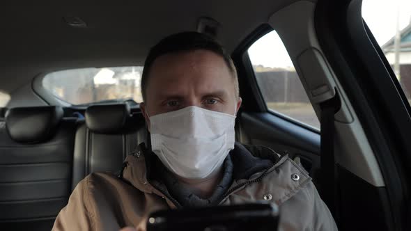 A Man in a Medical Mask Sits in the Car and Uses His Smartphone