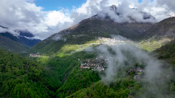 Clouds forming above scenic village in lush Italian alp valley hillside; drone alt