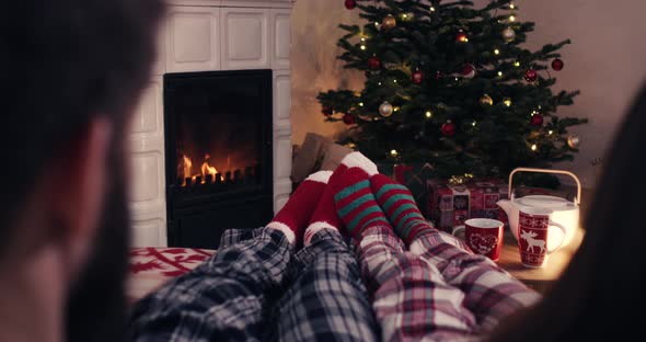 Over shoulder couple feet in christmas woolen socks near fireplace decorated tree tee cup in backg alt