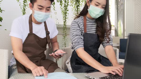 Asian couple in masks doing business calculations of restaurant and checking bills alt