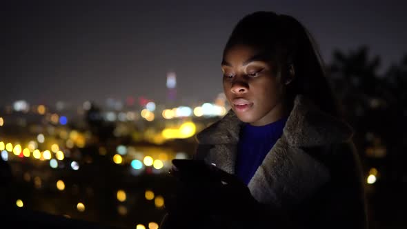 A Young Black Woman Works on a Smartphone and Smiles at the Camera in an Urban Area at Night alt