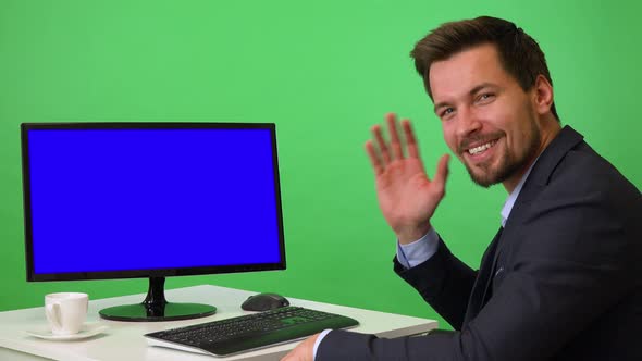 A Young Businessman Sits in Front of a Computer and Waves at the Camera with a Smile - Green Screen alt