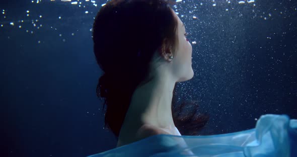 Portrait of a Brunette with Long Flowing Hair and a Blue Dress Under Water. She Turns, Puts Her Hand alt