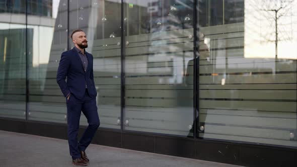 a Moustached and Bearded Man in a Blue Suit Has Hands in Pockets and is Walking Along the Glass Wall alt