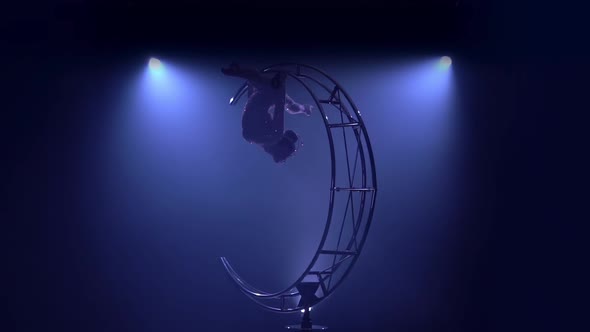 Gymnast Hangs Upside Down in a Twine on a Rotating Structure Moon. Blue Smoke Background. Slow alt
