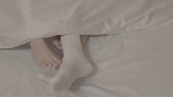 Man legs in white socks touched by two women with bare feet on bed on white bed sheets blanket close alt