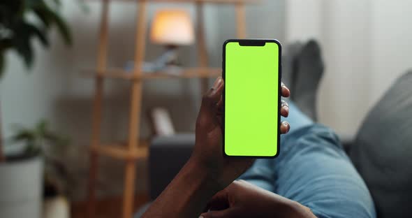 Young Man Looking at Mock Up Smartphone Screen While Lying on Sofa. Guy Using Phone While Watching alt