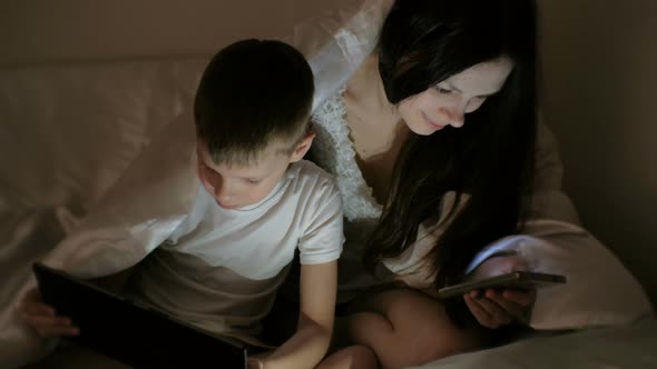Young Beautiful Woman Mom Browsing Internet in Her Phone While Her Son Watching Cartoon on Tablet
