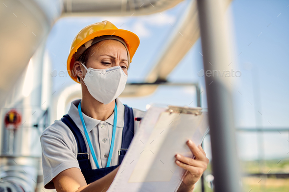 Female engineer wit face mask writing notes while inspecting outdoors industrial facility.